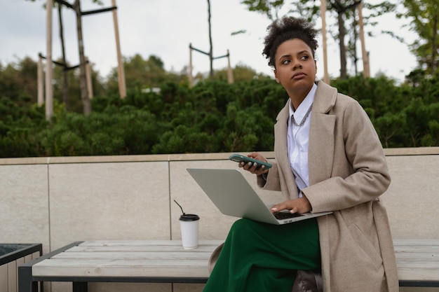 Afro-Amerikaanse jonge zakenvrouw die op een laptop werkt met een telefoon in haar handen, geen kantoor