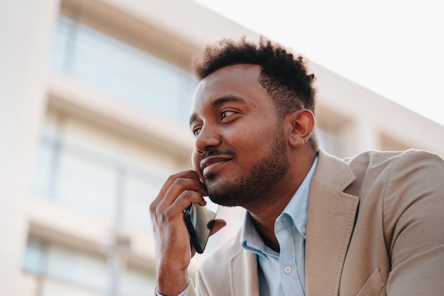 Afro-Amerikaanse jongen in een pak die op een smartphone praat met een klant van zijn bedrijf op een plek waar bedrijfsgebouwen te zien zijn. gesloten vlak.