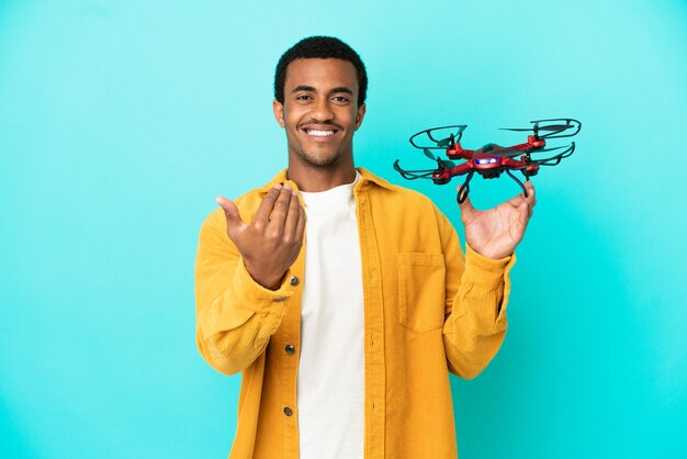 Afro-Amerikaanse knappe man met een drone over geïsoleerde blauwe achtergrond die uitnodigt om met de hand te komen. Blij dat je gekomen bent