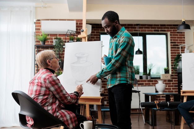 Afro-amerikaanse kunstleraar die tekenillustratie uitlegt aan senior vrouw die samenwerkt aan grafische schets in creatvity studio. divers team dat vaasmodel op canvas trekt en nieuwe artistieke vaardigheden leert