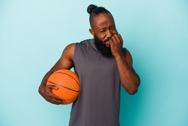 Afro-Amerikaanse man die basketbal speelt geïsoleerd op een blauwe achtergrond, vingernagels bijt, nerveus en erg angstig.