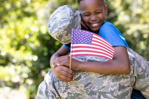 Afro-Amerikaanse man die een militair uniform draagt, naar huis terugkeert, zijn zoon omhoog houdt, omhelst, naar de camera kijkt en glimlacht, een jongen houdt een minivlag vast, op een zonnige dag