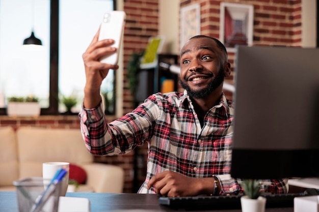 Afro-Amerikaanse man die foto's maakt met smartphone, met behulp van mobiele telefoon om foto's te maken thuis bureau. Werken op afstand als freelancer en online website gebruiken, werken aan startende ondernemingen.