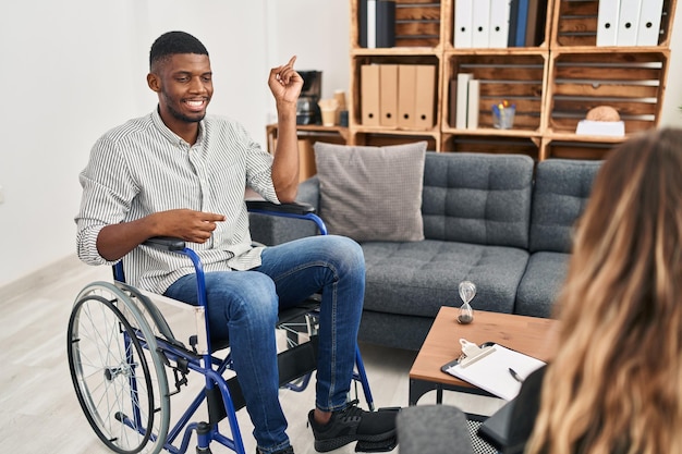 Afro-amerikaanse man die therapie doet zittend op een rolstoel met een grote glimlach op het gezicht wijzend met hand en vinger naar de zijkant kijkend naar de camera
