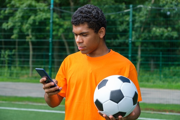 Afro-Amerikaanse man die voetbal vasthoudt en smartphone gebruikt op het sportveld