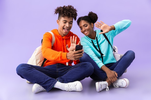 Afro-amerikaanse man en vrouw die rugzakken dragen die smartphone gebruiken terwijl ze op de vloer zitten met gekruiste benen, geïsoleerd over violette muur
