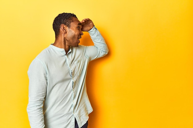 Foto afro-amerikaanse man in blauw shirt gele studio die ver weg kijkt met zijn hand op zijn voorhoofd