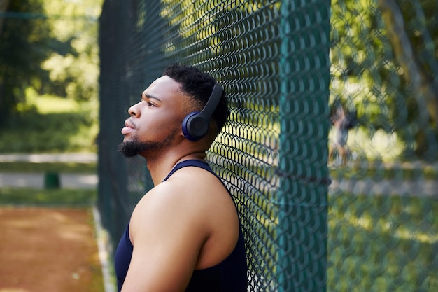Afro-Amerikaanse man in draadloze koptelefoon leunt op het metalen gaas van sportieve rechtbank en neemt pauze