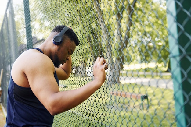 Afro-Amerikaanse man in draadloze koptelefoon leunt op het metalen gaas van sportieve rechtbank en neemt pauze