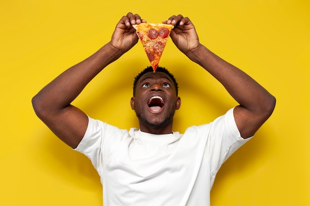 Afro-Amerikaanse man in wit t-shirt houdt een stuk pizza vast en opent zijn mond op gele achtergrond