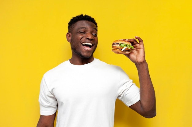 Afro-amerikaanse man in wit t-shirt houdt grote hamburger vast en verheugt zich op gele geïsoleerde achtergrond