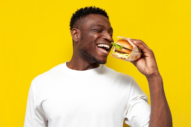 Afro-Amerikaanse man in wit t-shirt met grote hamburger en glimlachend, de man eet fastfood