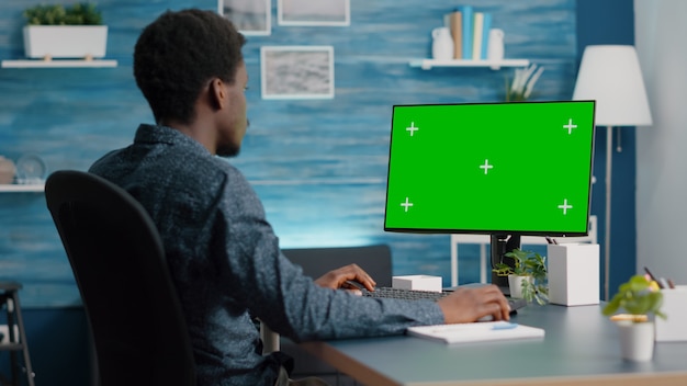 Afro-Amerikaanse man met behulp van en typen op mockup computer met groen scherm. Computergebruiker op geïsoleerde chroma mock-up display in woonkamer, licht huis