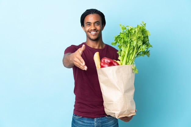 Afro-Amerikaanse man met een boodschappentas geïsoleerd op blauwe achtergrond handen schudden voor het sluiten van een goede deal