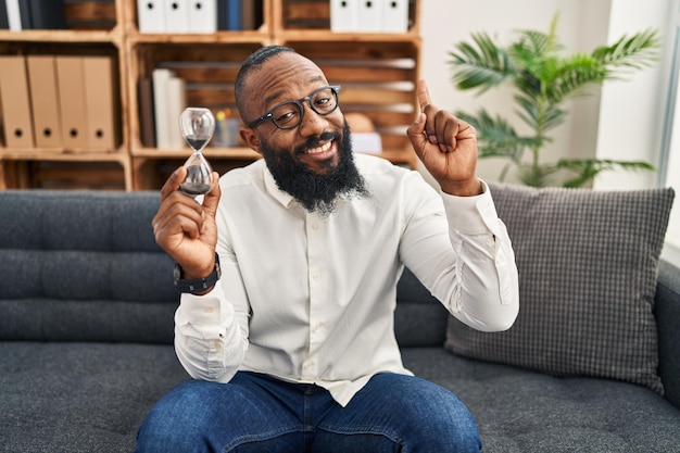 Afro-amerikaanse man met zandklok op kantoor glimlachend met een idee of vraag wijzende vinger met blij gezicht nummer één