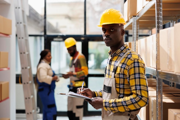 Afro-Amerikaanse man schrijft op klembord in industrieel pakhuisportret. Postkantoor pakhuis pakketbehandelaar draagt een helm met behulp van de checklist voor bestellingen en kijkt naar de camera