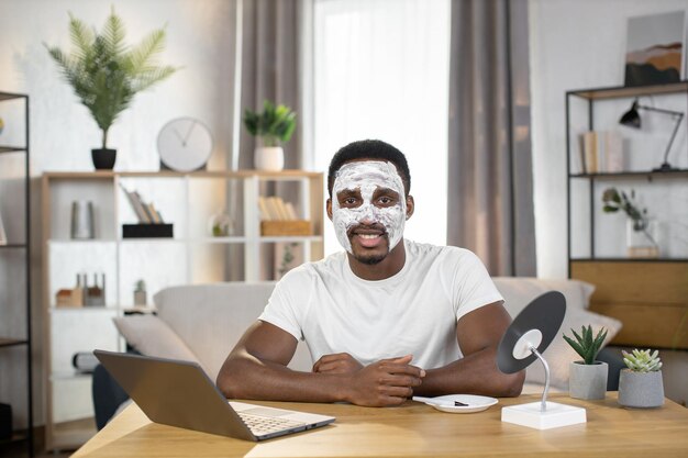 Afro-Amerikaanse man zit aan tafel met laptop thuis met gezichtskleimasker op zijn gezicht