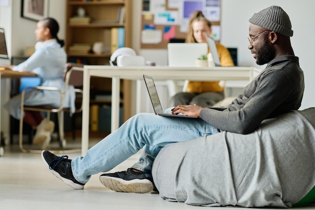 Afro-Amerikaanse man zit op een comfortabele zakstoel en werkt online op een laptop op kantoor