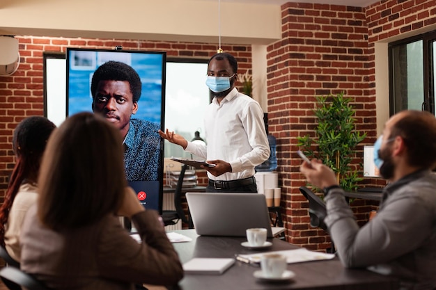 Afro-Amerikaanse manager met medisch gezichtsmasker tegen coronavirus die bedrijfsstrategie uitlegt aan teamwerk tijdens online videocall-vergaderingsconferentie in startup-kantoor. Zakelijk gesprek op monitor