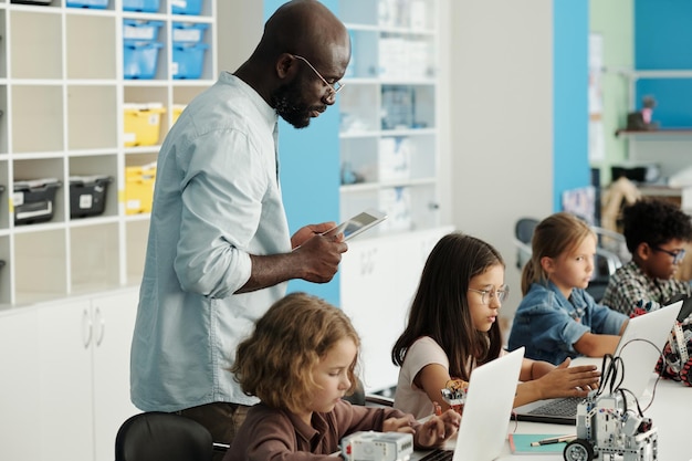 Afro-amerikaanse mannelijke leraar robotica die taak van schoolkinderen controleert