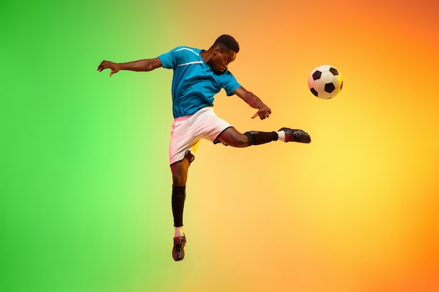 Afro-Amerikaanse mannelijke voetbal, voetballer training in actie geïsoleerd op gradiënt studio achtergrond in neonlicht. Concept van beweging, actie, prestaties, gezonde levensstijl. Jeugd Cultuur.