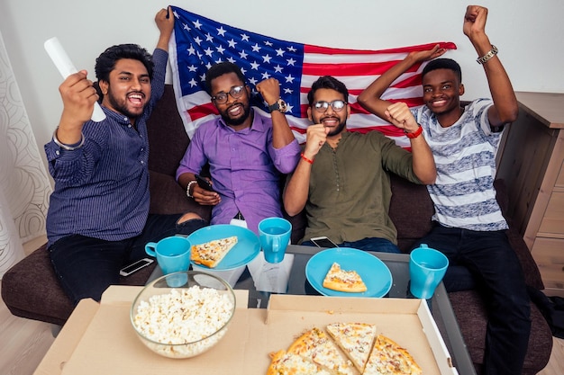 Afro-Amerikaanse mannen die pizza en popcorn eten, juichen en glimlachen terwijl ze thuis naar een tv-wedstrijd kijken