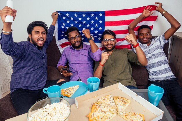 Afro-Amerikaanse mannen eten pizza en popcorn, juichen en glimlachen terwijl ze thuis tv-wedstrijd kijken.