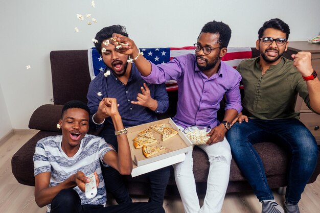 Afro-amerikaanse mannen eten pizza en popcorn, juichen en glimlachen terwijl ze thuis tv-wedstrijd kijken.