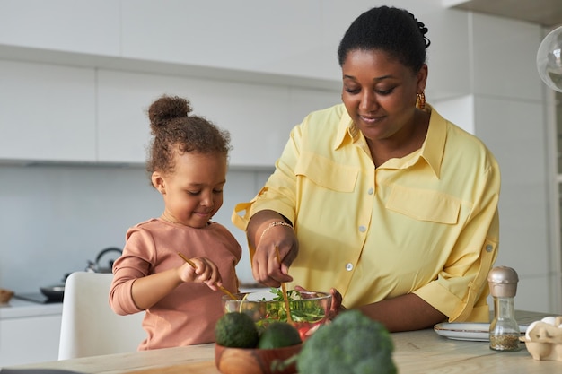 Afro-Amerikaanse moeder kookt groentesalade in de keuken met dochtertje dat haar helpt