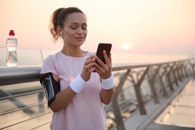Afro-Amerikaanse mooie sportvrouw in roze t-shirt en oortelefoons, met smartphonehouder, met behulp van mobiele telefoon tijdens het sporten bij zonsopgang, browsen door mobiele apps, staande op de loopband van de stadsbrug