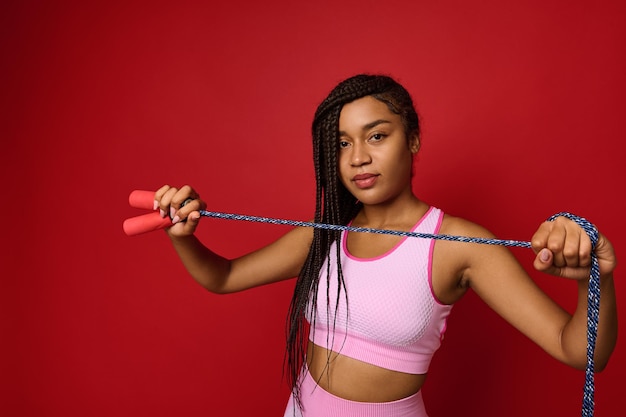 Afro-Amerikaanse mooie vrouw in roze sportkleding houdt een springtouw voor haar en toont het aan een camera die er zelfverzekerd naar kijkt, poserend over rode achtergrond met kopieerruimte voor reclame