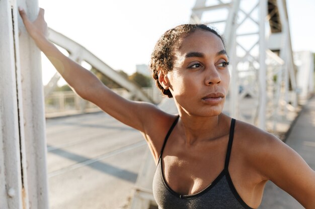 Afro-Amerikaanse mooie vrouw in sportkleding die opzij kijkt tijdens het trainen op de oude brug