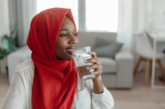 Afro-Amerikaanse moslimvrouw die vers mineraalwater uit glas drinkt en opzij kijkt naar vrije ruimte