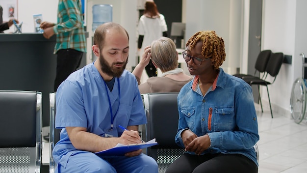 Afro-Amerikaanse patiënt praat met verpleger over medische ondersteuning en behandeling om de diagnose te genezen, chatten in de wachtkamer van de ziekenhuisreceptie. Mensen doen overleg in de lobby.