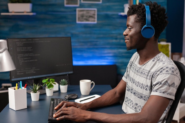 Afro-Amerikaanse programmeur met koptelefoon die programmacode schrijft op computer die op afstand van huis werkt. Werknemer man codering zakelijke beveiligingsserver. Concept van softwareontwikkeling