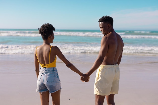 Afro-amerikaanse shirtloze jonge man die de handen van de vrouw vasthoudt terwijl hij op het strand tegen de hemel staat