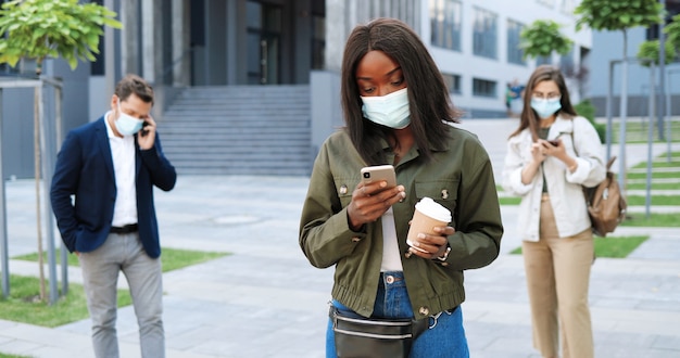 Afro-Amerikaanse stijlvolle jongedame in medische masker staande op straat en SMS-bericht op smartphone. Vrouw buiten op mobiele telefoon te tikken en koffie te houden. Pandemie. Mensen van gemengde rassen.