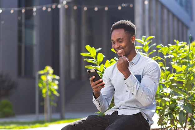 Afro-Amerikaanse student man succesvol leest de resultaten van zijn werk online vanaf de telefoon, buitenshuis, glimlacht en verheugt zich