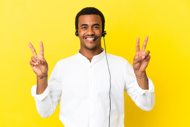 Afro-Amerikaanse telemarketeer man aan het werk met een headset over geïsoleerde gele achtergrond met overwinningsteken met beide handen