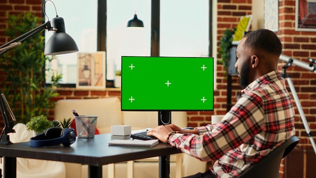 Afro-Amerikaanse telewerkende freelancer die gegevens invoert op chroma-toets mock-up computer groen scherm in de woonkamer van het appartement Ontspannen externe werknemer die taken uitvoert terwijl hij thuis werkt