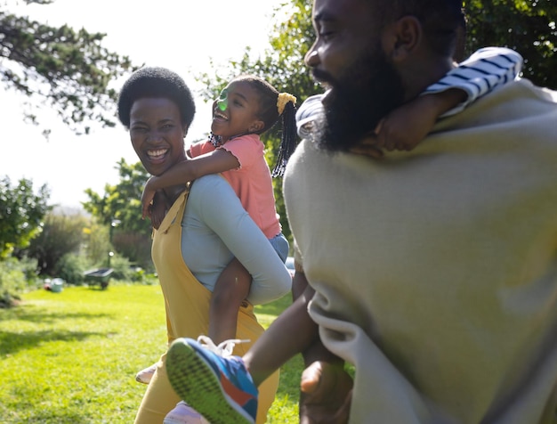 Afro-Amerikaanse vrolijke ouders dragen hun zoon en dochter op hun rug terwijl ze in de tuin lopen. Ongewijzigd, levensstijl, familie, liefde, saamhorigheid, jeugd, spelen en plezier concept.