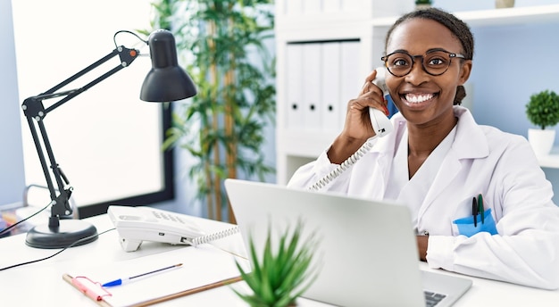 Afro-Amerikaanse vrouw arts met behulp van laptop praten aan de telefoon in de kliniek