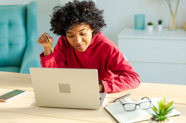 Afro-Amerikaanse vrouw die laptop gebruikt die online winkelt en betaalt met een gouden creditcardmeisje dat thuis zit