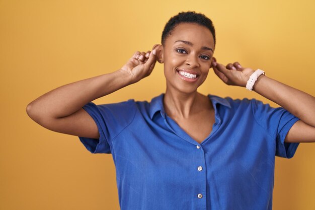 Afro-Amerikaanse vrouw die over een gele achtergrond staat en glimlacht en oren trekt met vingers, grappig gebaar. auditie probleem