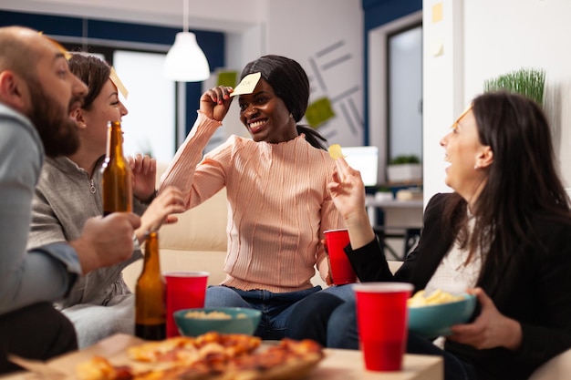 Afro-Amerikaanse vrouw die raadt wie met collega's speelt, viert met een drankje na het werk. Kantoorcollega's genieten van charades pantomime spelen met plaknotities op een feestje.