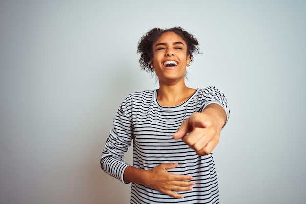 Afro-Amerikaanse vrouw, gekleed in een marineblauw gestreepte t-shirt die over een geïsoleerde witte achtergrond staat en naar je lacht, wijzende vinger naar de camera met hand over lichaamschaamte-expressie