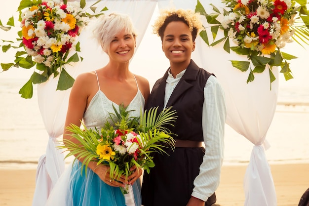 Afro-Amerikaanse vrouw in de rol van bruidegom en kortblonde bruid in blauwe jurk in ceremonie onder huwelijksboog op tropisch strand