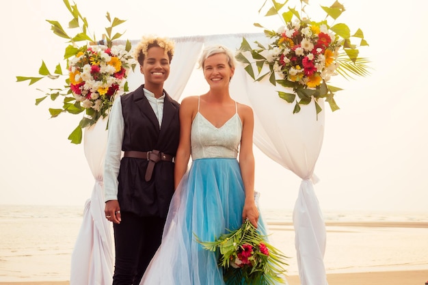 Afro-Amerikaanse vrouw in de rol van bruidegom en kortblonde bruid in blauwe jurk in ceremonie onder huwelijksboog op tropisch strand