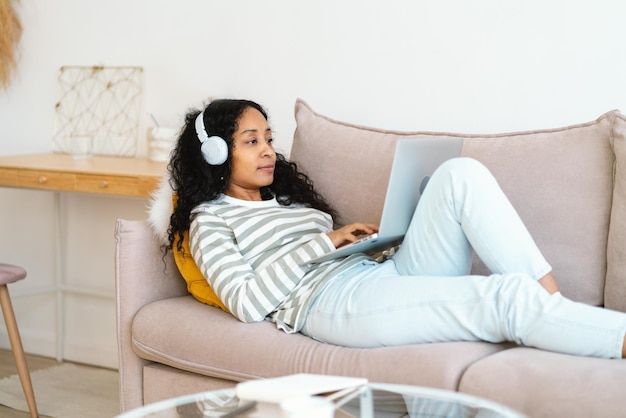 Afro-Amerikaanse vrouw in headset die online op laptop chat terwijl ze op de bank in de woonkamer ligt