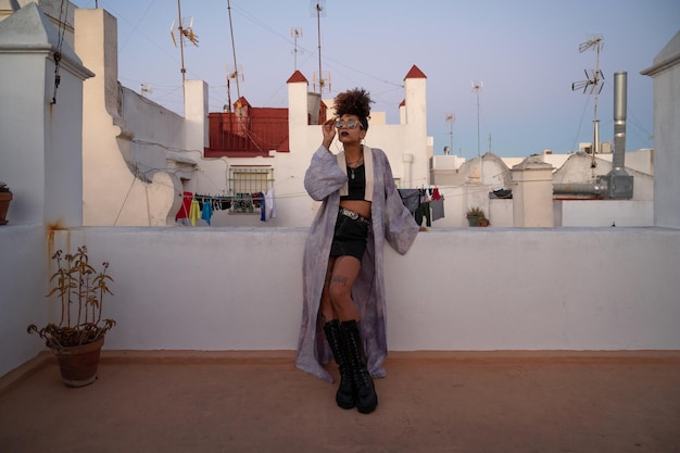 Afro-Amerikaanse vrouw in stijlvolle kleding die een zonnebril aanpast en op een barrière tegen huizen leunt terwijl ze 's avonds op het terras ontspant in Cadiz, Spanje?